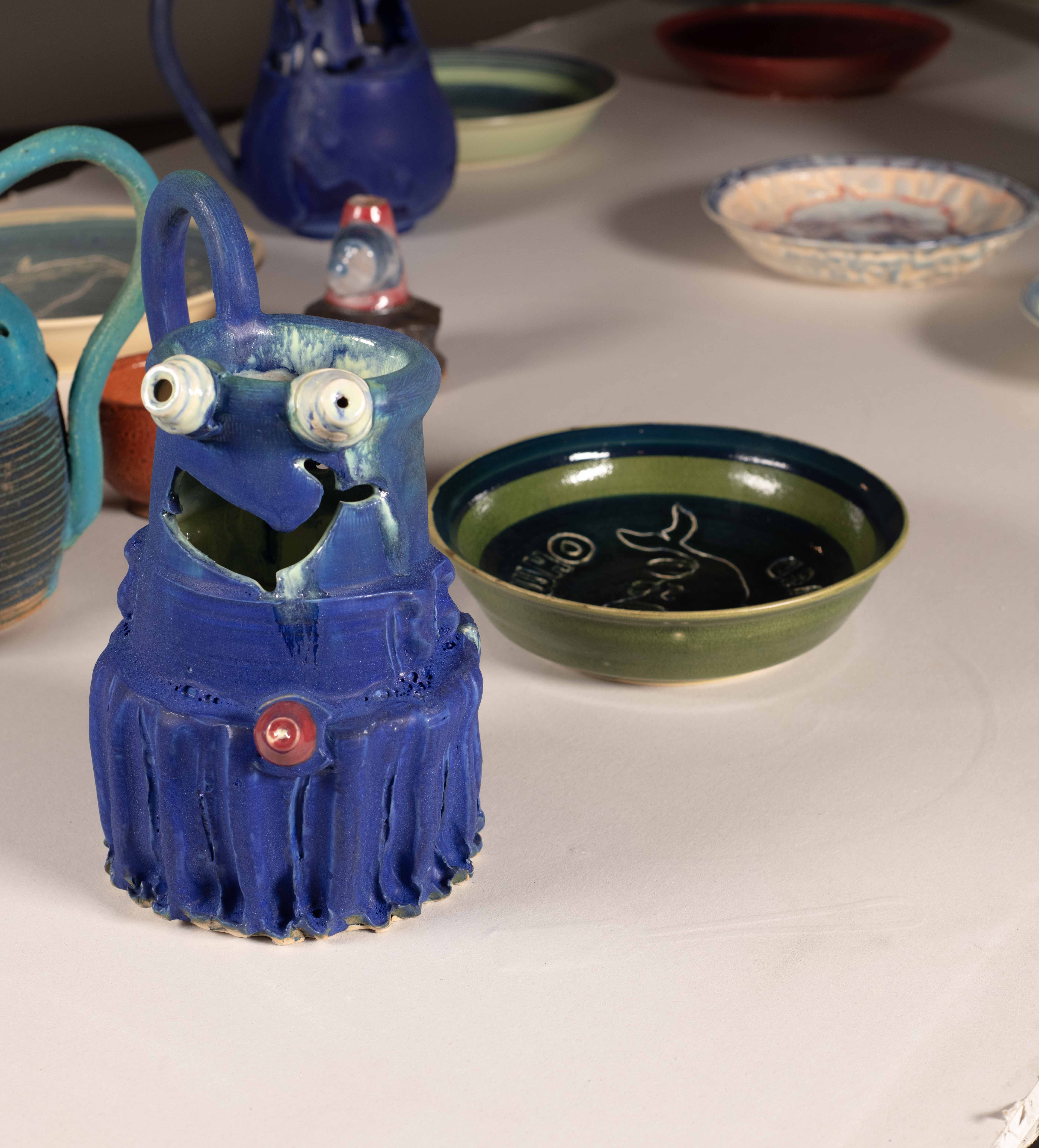 Drywall table with LED lights imbedded below the surface. 25 pound iron weights for  lifting are the tables wheels. The table legs are an outline of a horses feet. On top of the table  are an assorted gathering of brightly glazed Ceramic objects— Puzzle Jugs and WHO plates.