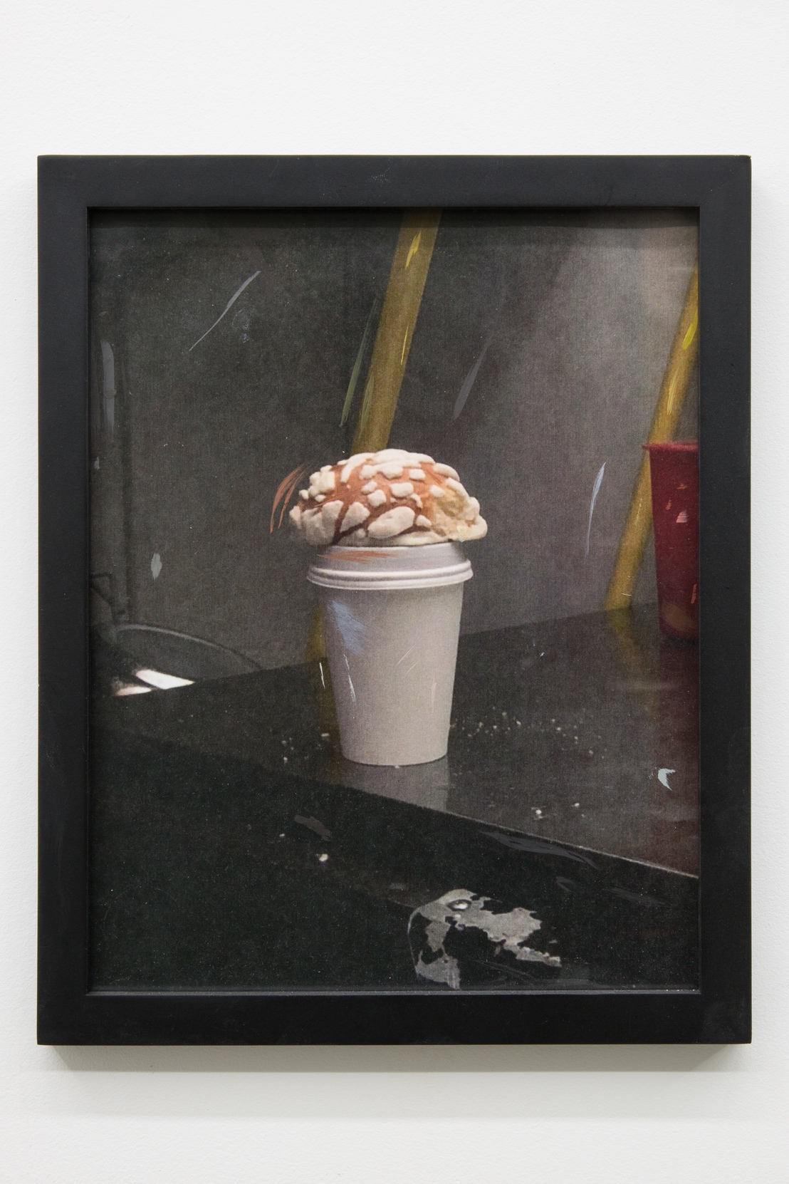 Detail image of a printed photograph framed in a thin matte black frame. The printed image has small marks of oil paint. The image is of a concha pastry sitting on top of an unlabeled coffee cup. The coffee cup is placed at the edge of a stainless steel work fridge. In the background is the corner of a sanitizing bucket and the bottom of a metal champagne bucket.