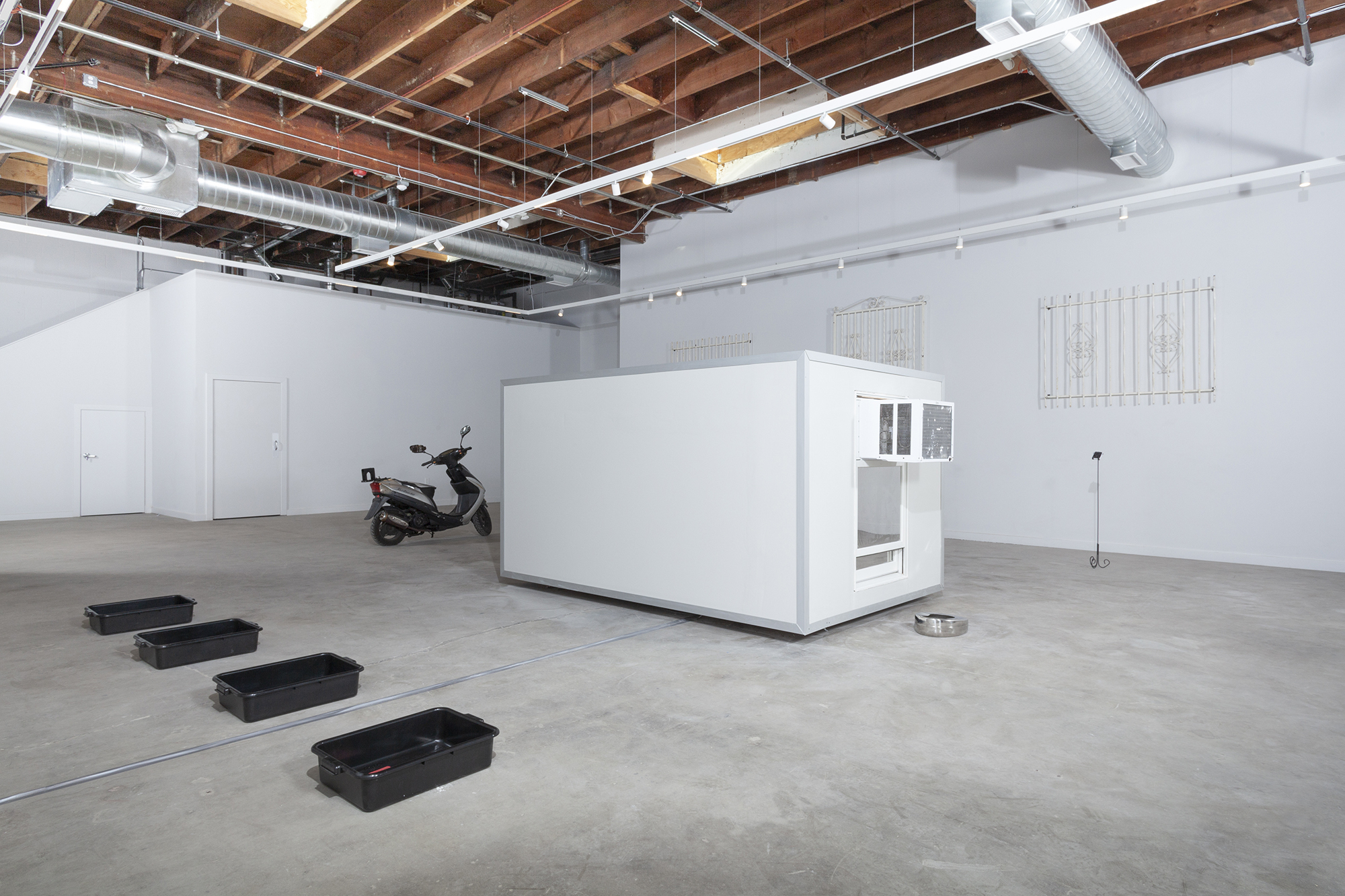 Installation image of Hello Baby, Terroir, an exhibition at Murmurs Gallery in Los Angeles. The picture is shot at a wide angle. The room is white with gray concrete floors and exposed wooden beams along the high ceilings. At the center of the room is a built structure that appears to hover several inches off the ground. Behind it is a parked scooter. The parked scooter contains a thick stack of research documents made inaccessible through being sealed underneath plexiglass in the storage seat of the scooter. To the left of it are four black TSA bins placed on the floor. There are three ornate white metal grates installed midway up the wall. In front of each window is a freestanding curvilinear stand which holds separate baby monitors. Each baby monitor corresponds to a camera on the inside of the center structure and surveils the sculptures within. There are two windows treated with privacy film on either side, each with its own functioning air conditioner. Underneath each air conditioner is a bed pan filled with non-domestic coins and water. The door to the structure is located on the ceiling, out of reach to all viewers. The only way into the structure is through a dog door built into said out-of-reach door.