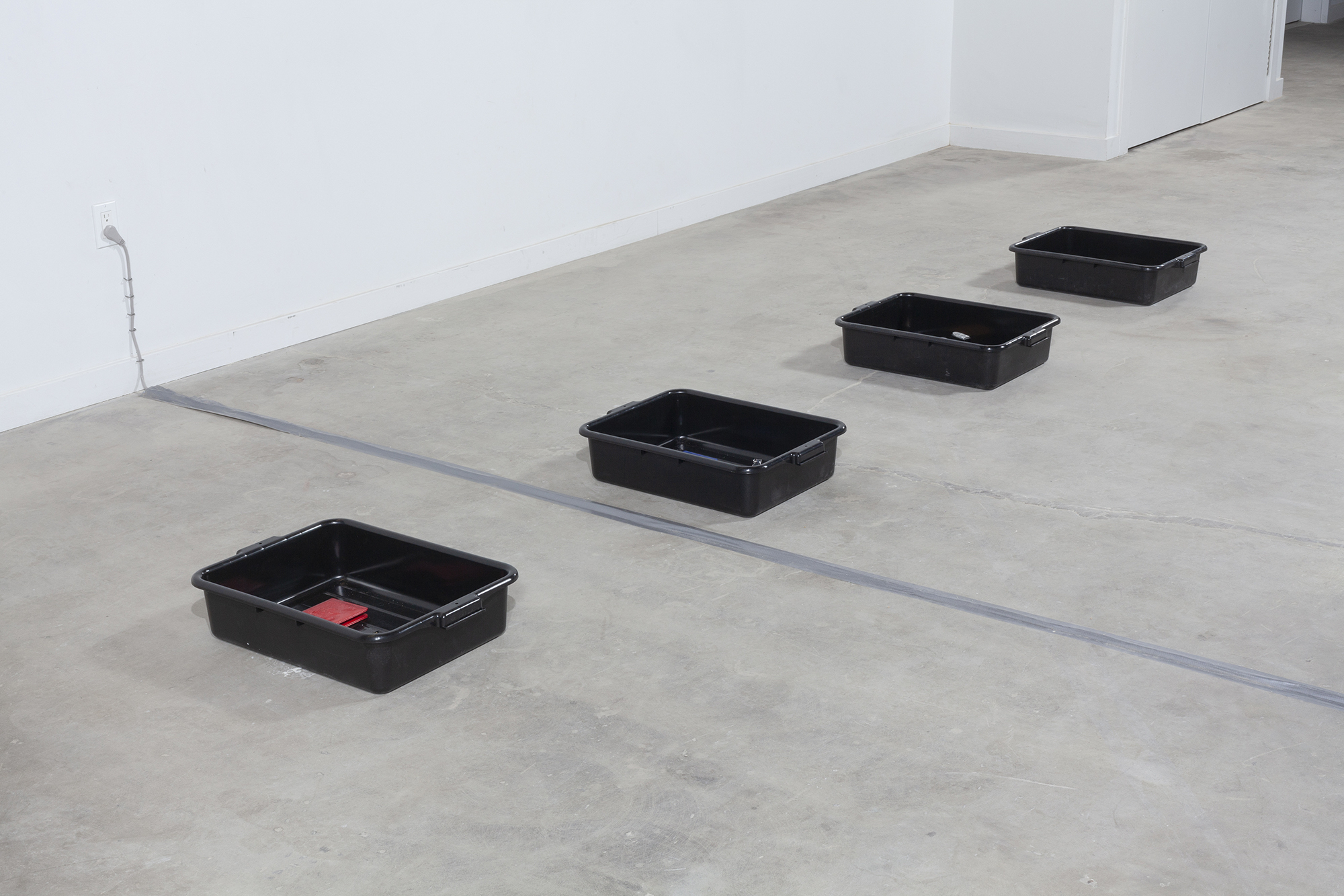 Installation image of four black TSA bins sitting on the floor filled with different compositions of objects.