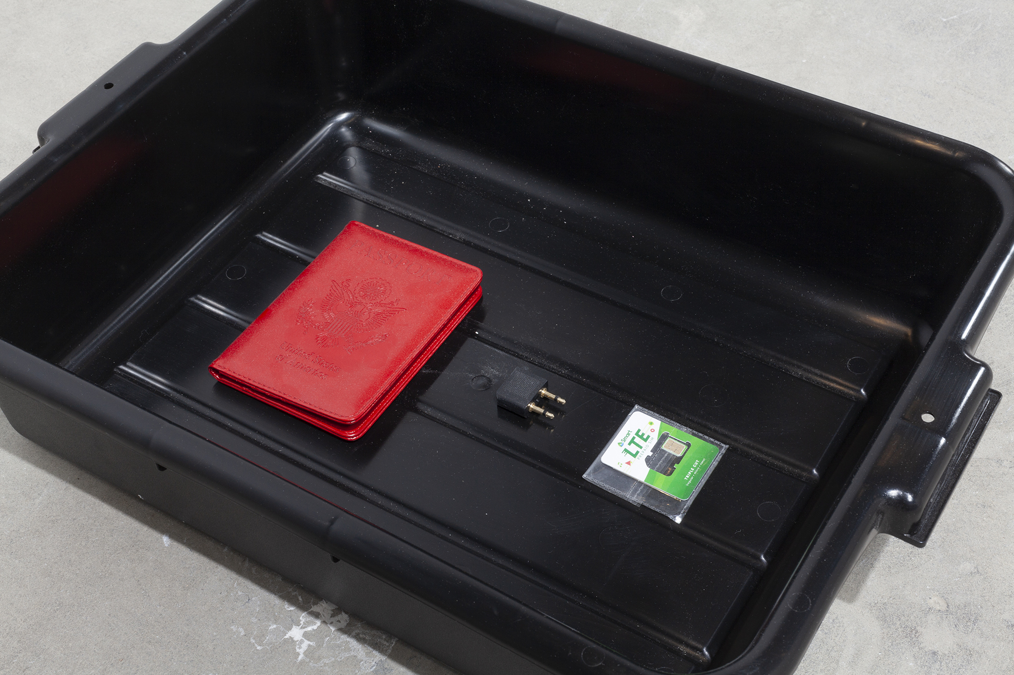 Detail image of a black TSA bin sitting on the floor. Inside of it are the following items: a red American passport holder, a foreign SIM card, a needle, and an airplane headphone adaptor.