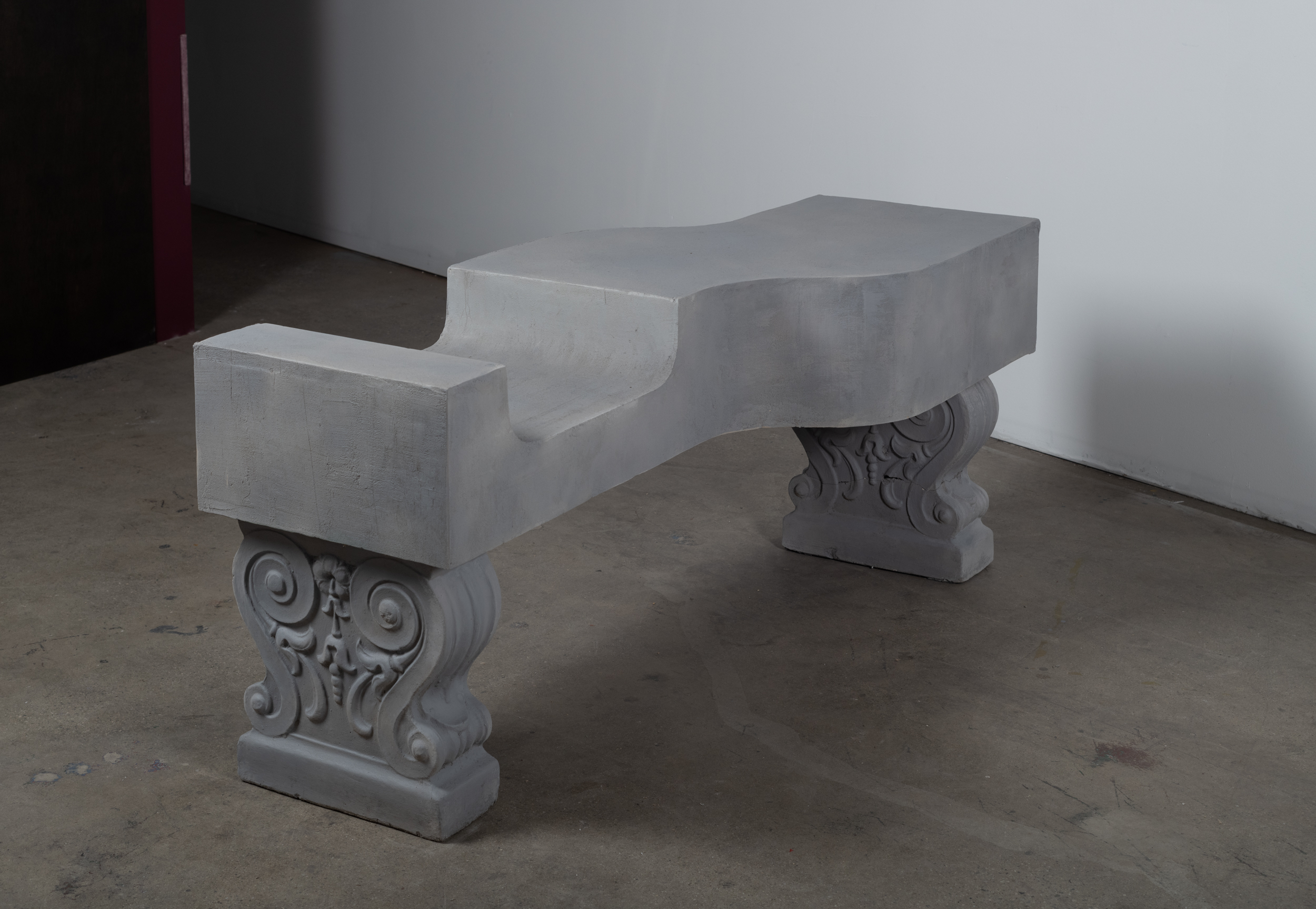 This image is a detail shot of an undulating concrete bench. The bench itself rests on top of two ornate bases. There is a one ergonomic dip along the seated portion of the bench which tells the viewer where to place their body.